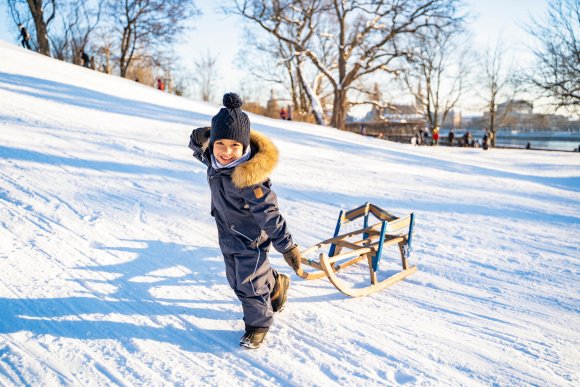 African American or Latin boy in warm overalls is pulling handmade wooden sled